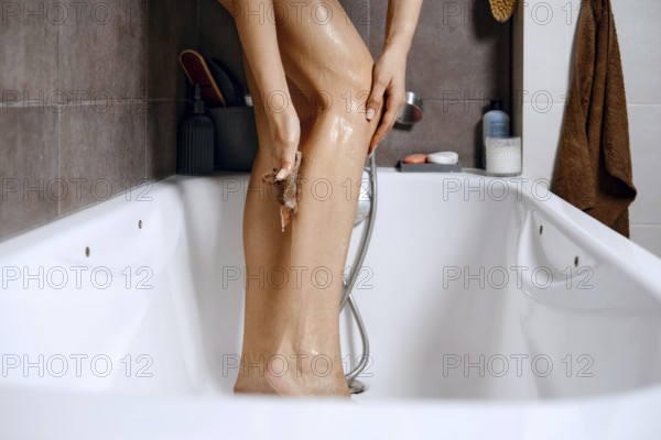 A woman stands in a bathtub. She uses a scrubbing tool to wash her leg. The bathroom has tiles, a