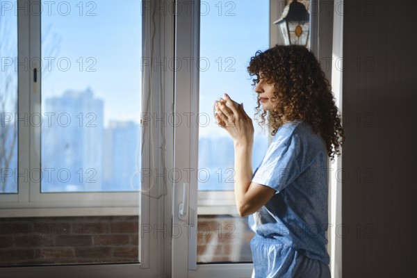 A woman stands by an opened for ventilation balcony door in the morning, looking outside while