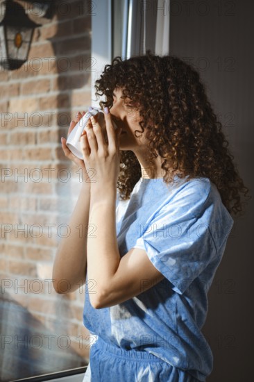 A woman stands by a window and holds a warm drink close to her face. Sunlight shines through the