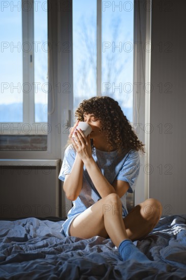 A woman sits on a bed with a blue blanket, holding a white cup. Bright winter sunlight comes