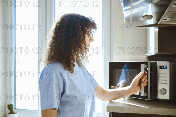 A woman stands by a microwave in a kitchen. She is preparing to heat food. Sunlight enters through