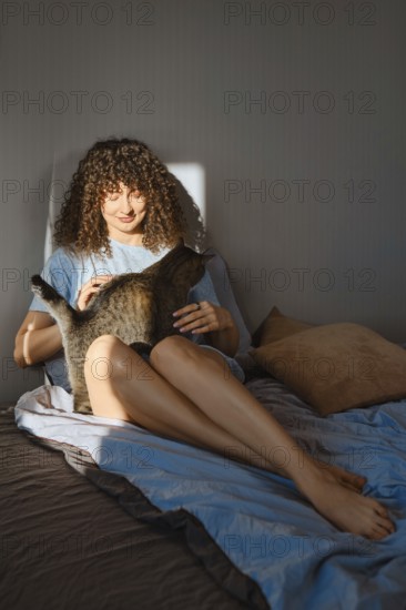 A woman sits on a bed in a cozy room during morning light. She gently pets a cat sitting on her lap