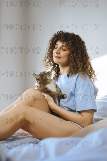 A woman sits on her bed with her cat in her lap. She looks happy and relaxed while sunlight shines