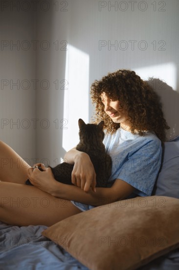 A woman with curly hair sits on a bed while holding a cat. Sunlight comes through the window,