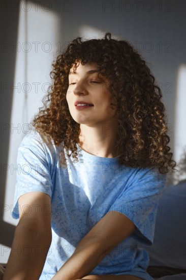 A woman with curly hair sits in a well-lit room. Sunlight streams through the window, creating warm