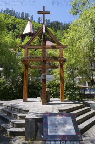 Wooden cross on a pedestal surrounded by green trees and a building, memorial to the tragedy of
