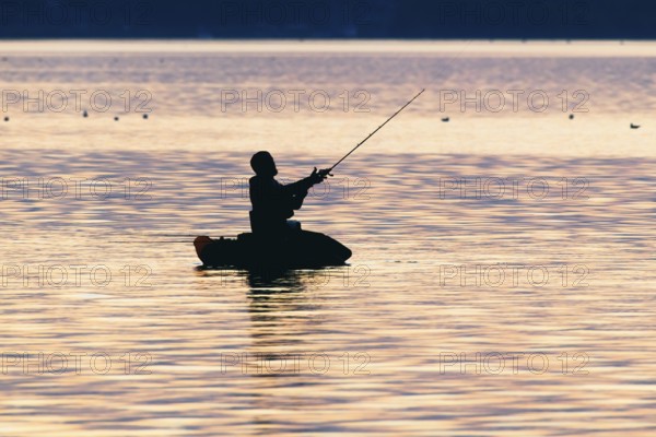 Angler sport fisherman in his belly boat in orange-red lake at sunset, silhouette in calm water,