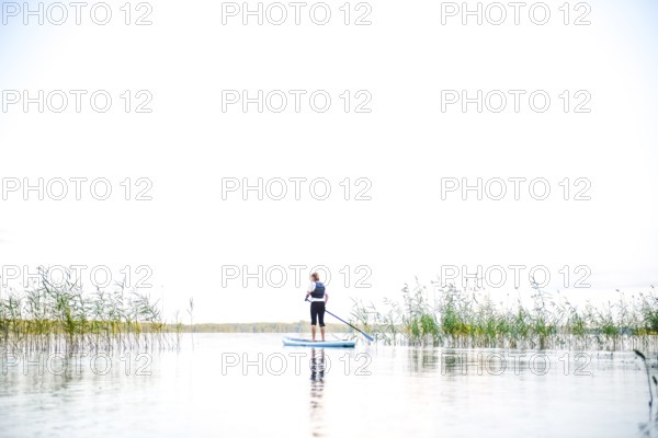 Person on a stand-up paddle board between reeds on a calm lake under clear skies, Asnen, Kronborgs