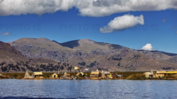 Row of reed huts on a calm lake under a partly cloudy sky and mountain backdrop, The floating reed