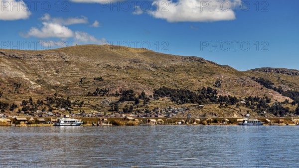 A calm lake in front of a mountainous landscape with boats and a village on the shore under a blue