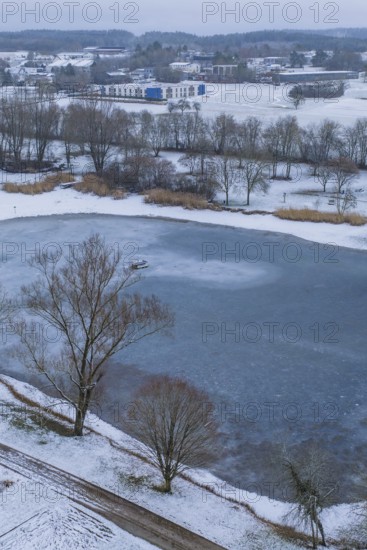 Small lake surrounded by snow-covered park and village in the background, icy surface, Empfingen,