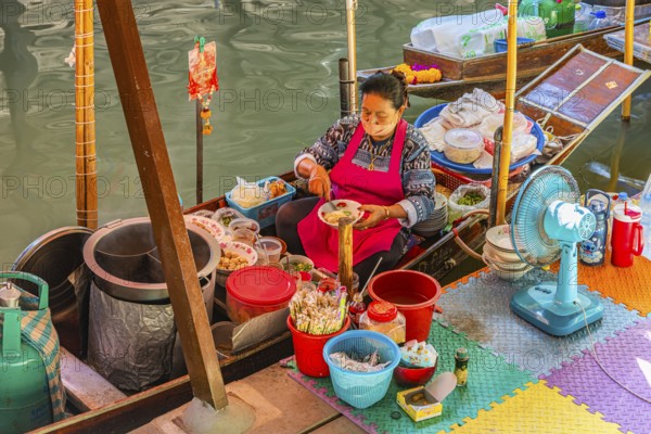 Elderly woman wearing mask offers food on her wooden boat, Damneon Saduak floating markets channel,