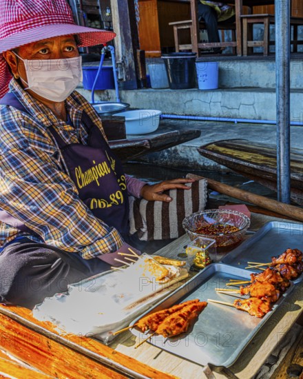 Elderly woman wearing mask sells barbecue skewers on her wooden boat on the Damneon Saduak floating