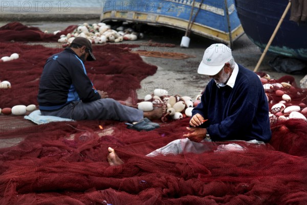 Fishermen repair nets in harbor surrounded by blue boats and tools, Essaouira, Marrakesh-Safi,