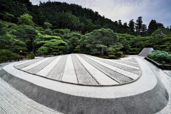 Zen garden of Ginkaku-ji Temple with precise sand patterns and rocks surrounded by green landscape,
