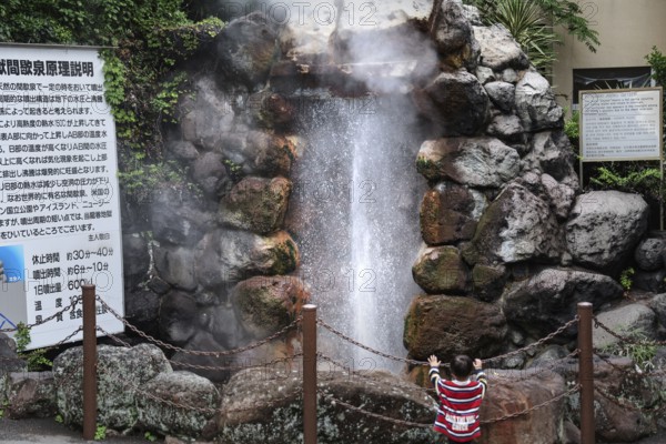 Steaming geyser surrounded by rocks and vegetation, with information board and barrier, Beppu,