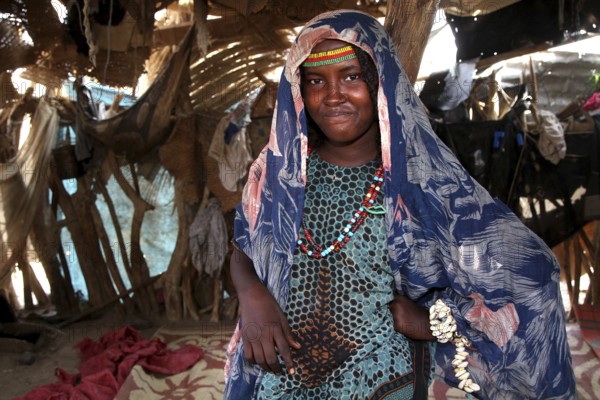 Aishe stands in Muhammad's hut wearing traditional clothes surrounded by coloured fabric in