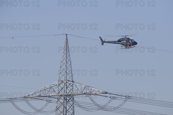 A helicopter carries out technical work on power lines next to a mast under a blue sky, an employee