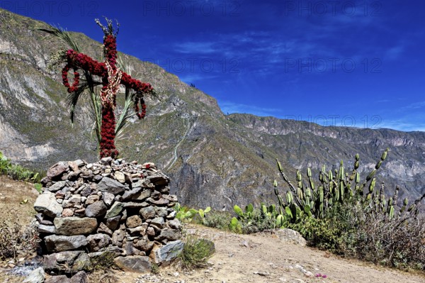 A cross of flowers and branches stands on a pile of rocks in a dry desert landscape with cacti,