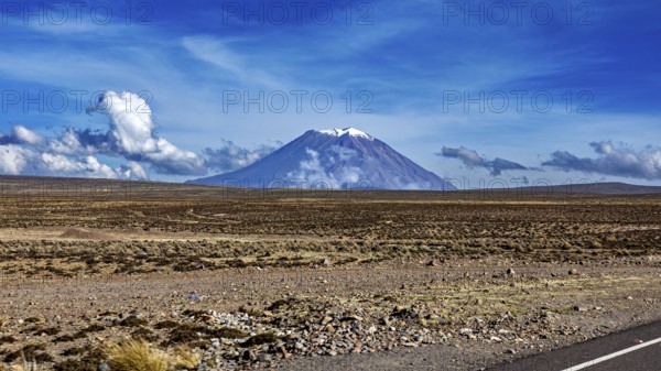 Volcano in a wide desert landscape under blue sky with clouds, snow-capped summit, The Andean