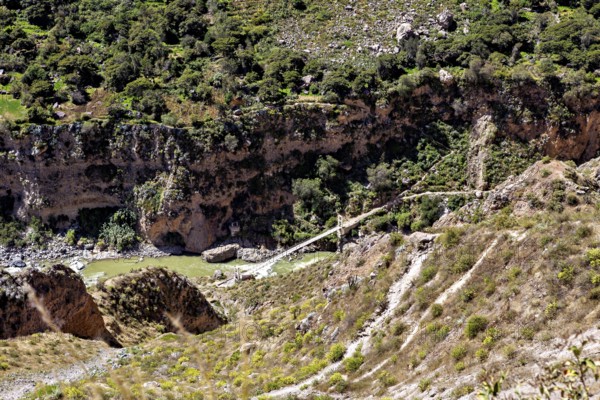Wooden bridge across river in an overgrown valley surrounded by rocks and thick greenery, The