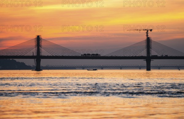 A boat sails on the river Brahmaputra during sunset, with the silhouette of a cable-stayed bridge
