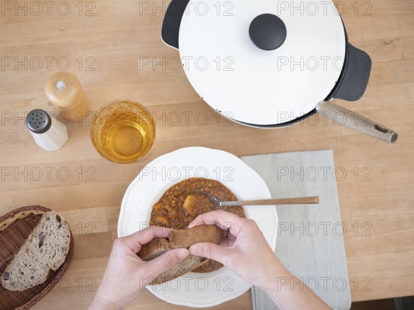 Hands breaking slice of bread over bowl of lentil soup, served with drink, salt and pepper, and