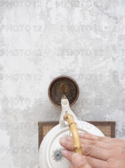 Hand gracefully pours tea from a white ceramic teapot with bamboo handle into a small brown bowl on