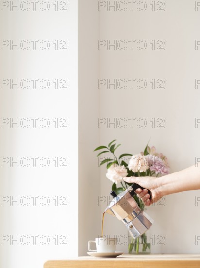 Hand pouring coffee from moka pot into cup on wooden table, with flowers in background, in