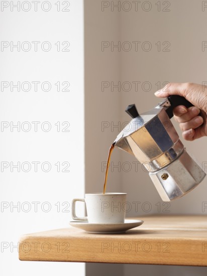 Hand pouring coffee from a moka pot into a cup, placed on a wooden table, creating a cozy and