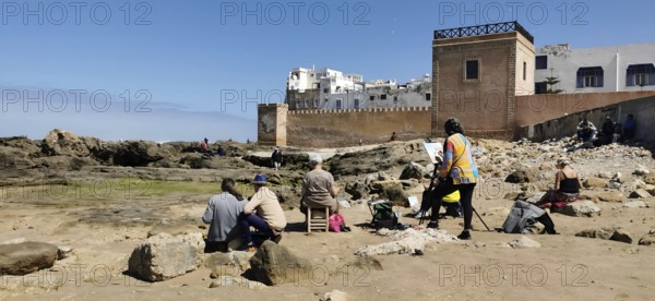 People sitting with canvas on the rocky beach of Essaouira the city view in front of the city wall