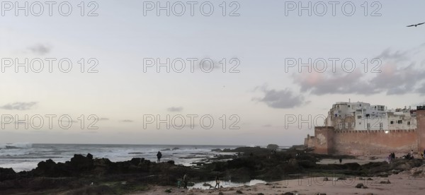 Evening on the coast with city walls of Essaouira, people standing on rocks and gentle waves at