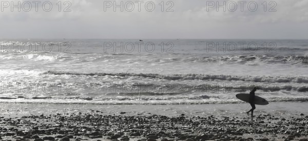 Silhouette of a surfer carrying his board along a pebble beach with a view of the wavy ocean, Sidi