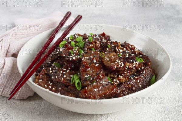 Crispy beef, with green onions and sesame seeds, in a bowl with chopsticks, on a textured surface,