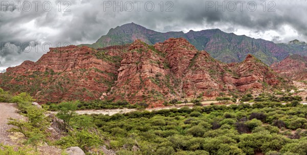 Red rock formations rise against green vegetation and cloudy skies, the landscape and colorful rock
