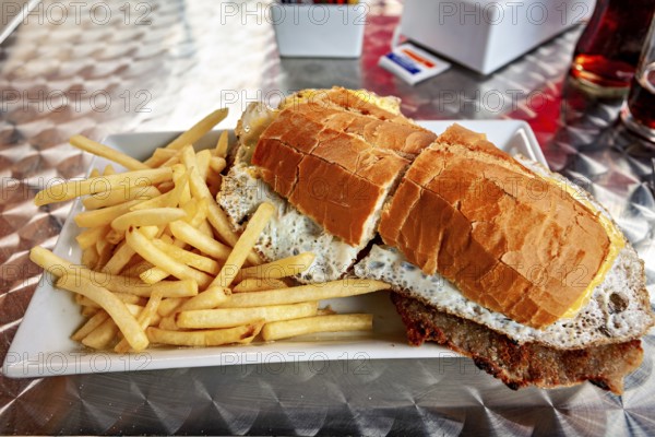 Large sandwich with fries served on a metal table, thickly filled with bread and a fried egg