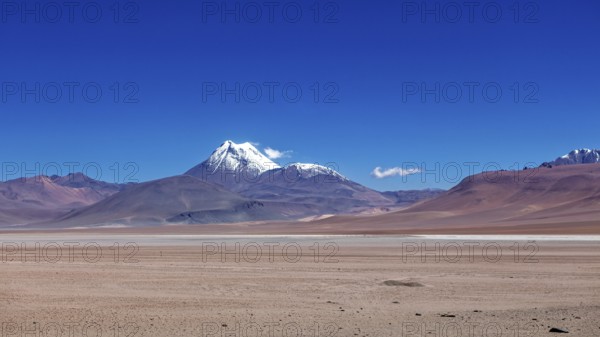 A snow-covered volcano rises above a dry, vast desert under clear skies, the landscape and colorful
