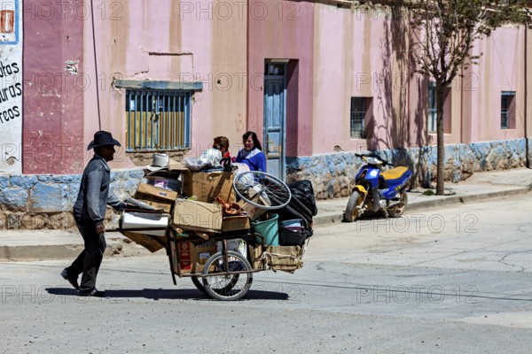 Man pulls carts full of packages past a colorful building, people in the streets of San Antonio de
