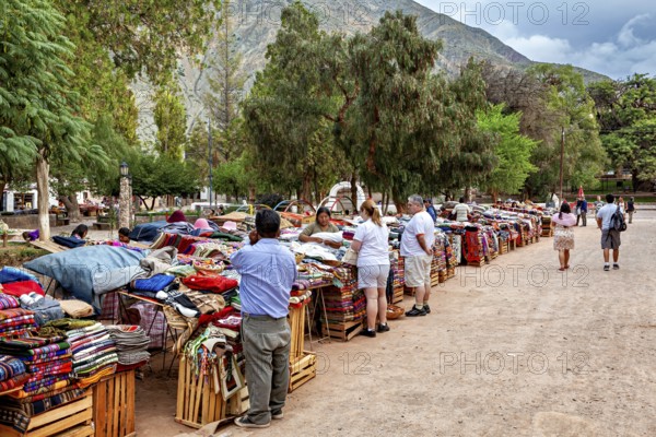 People check out textile stalls at a lively market against a mountain backdrop, tourist and