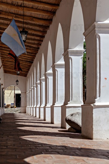 Shady arcade with white columns and wooden ceiling. An Argentinean flag is seen, The arcades of