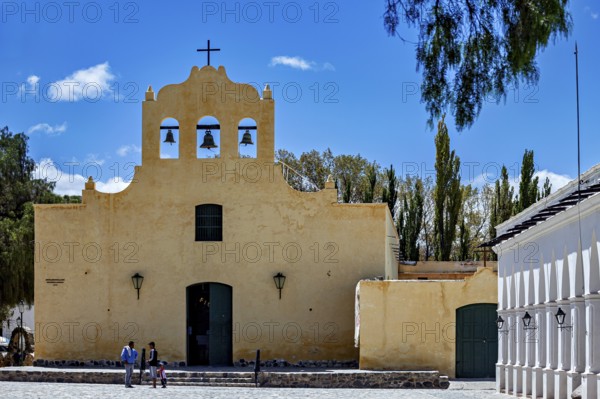 Yellow church with bell tower under blue sky, people on stairs surrounded by trees, Iglesia San