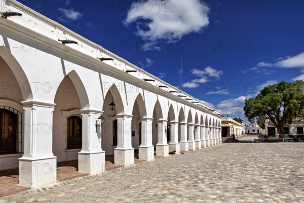 Arcades along a paved square in sunny weather with blue skies and trees, The arcades of Cachi in