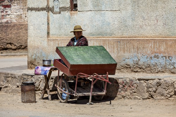 Woman wearing hat sitting at a cart with a green roof on the street corner, people in the streets