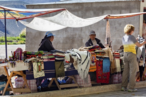 Two woman at market stalls surrounded by handmade blankets, interacting with a customer, tourist