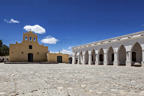 Square with yellow church and white arcades under bright blue sky, Iglesia San José de Cachi in