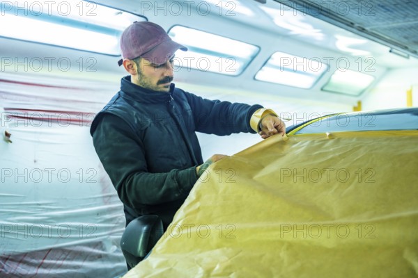 Auto body technician carefully applying masking paper and tape to a vehicle in a professional car