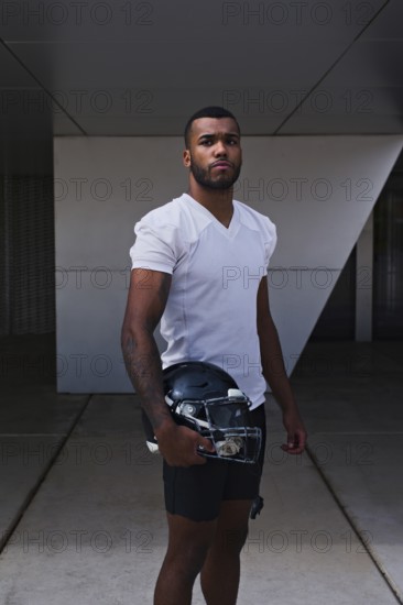 A confident American football player stands in a white jersey, holding a helmet, ready for the game