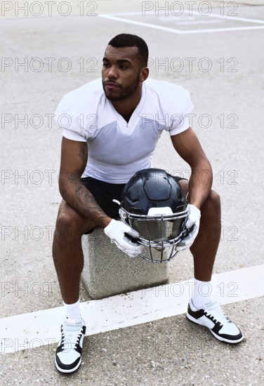 An american football player in a white jersey sits on a bench holding his helmet, reflecting before