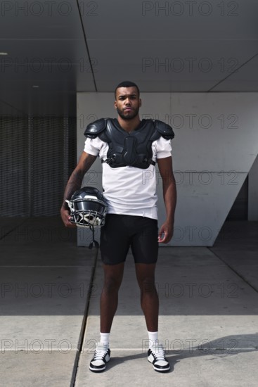 A determined American football player stands confidently, wearing shoulder pads and holding a