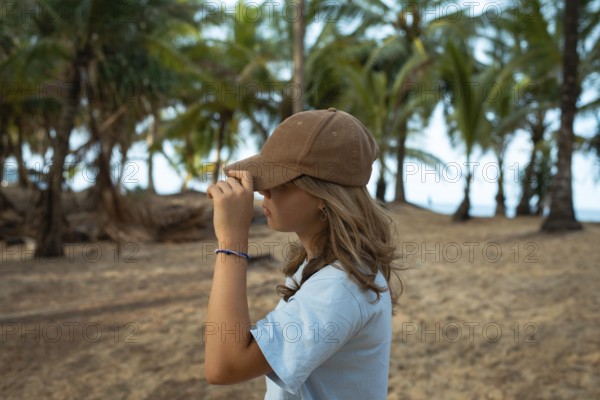 A young woman wearing a casual brown cap and blue shirt stands on a sandy beach, surrounded by lush